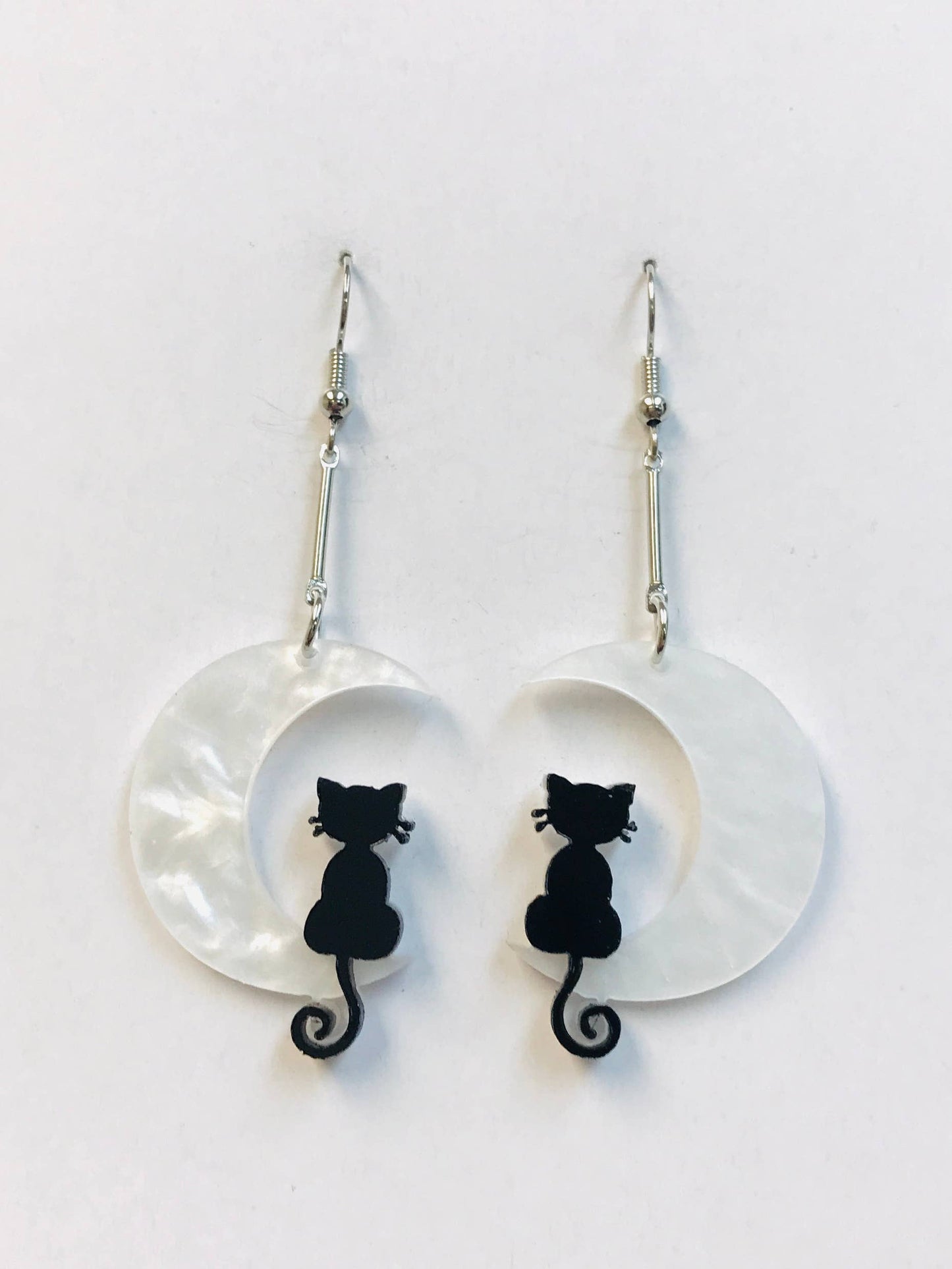 A pair of whimsical acrylic earrings featuring black cats sitting inside crescent moons. The lightweight earrings dangle from silver fish hook wires, offering a playful and mystical design perfect for cat lovers or Halloween outfits.