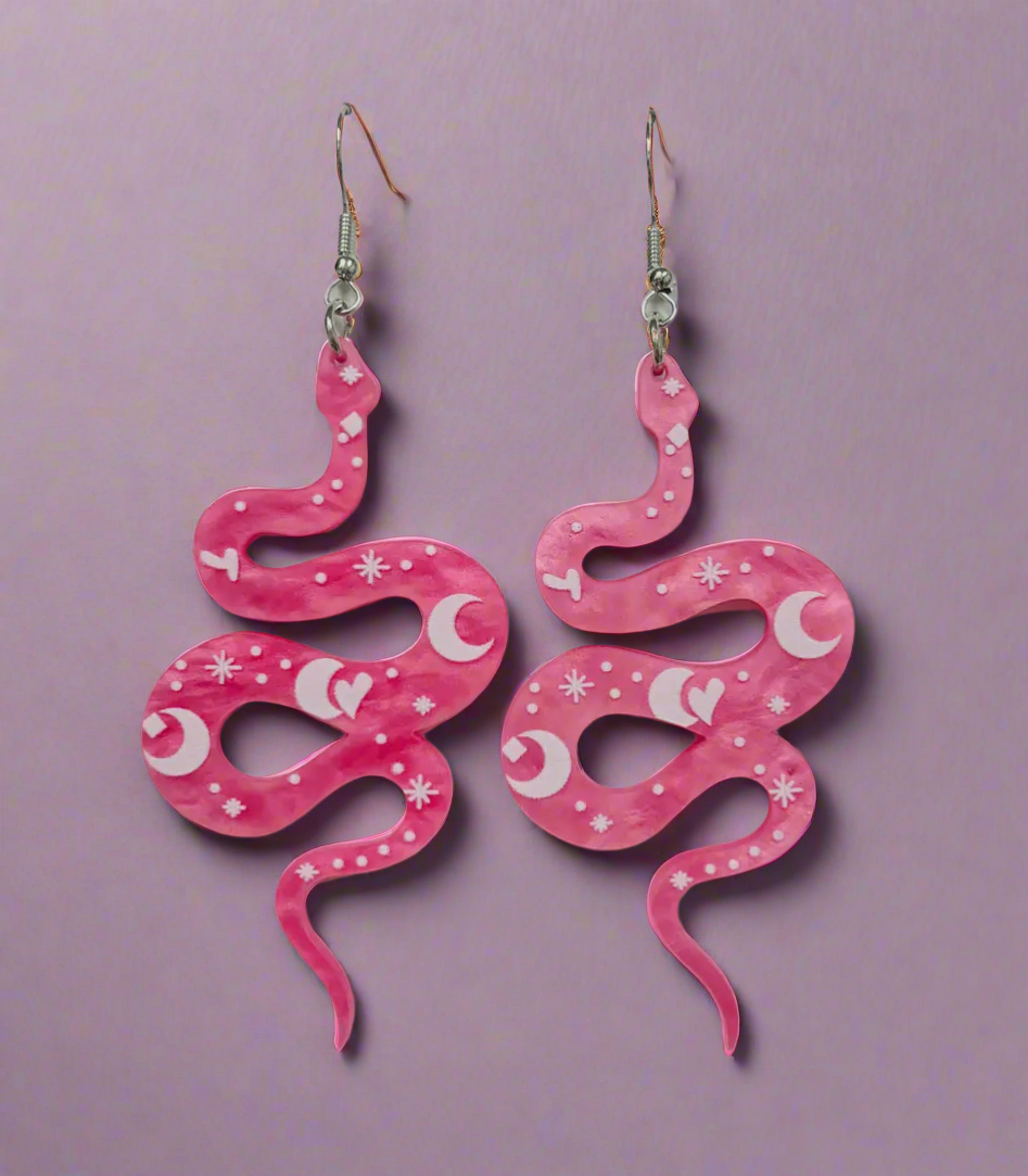 A pair of vibrant pink snake-shaped acrylic earrings featuring whimsical crescent moon and star designs. The lightweight earrings dangle from silver fish hook wires, combining a playful mix of celestial and snake motifs for a bold, statement-making accessory.