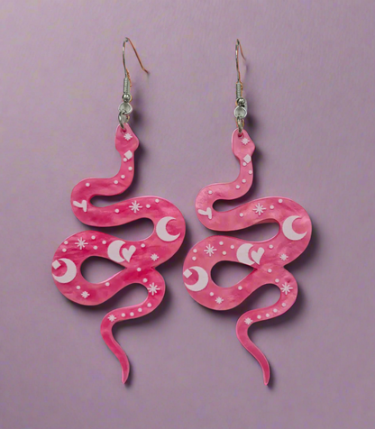 A pair of vibrant pink snake-shaped acrylic earrings featuring whimsical crescent moon and star designs. The lightweight earrings dangle from silver fish hook wires, combining a playful mix of celestial and snake motifs for a bold, statement-making accessory.