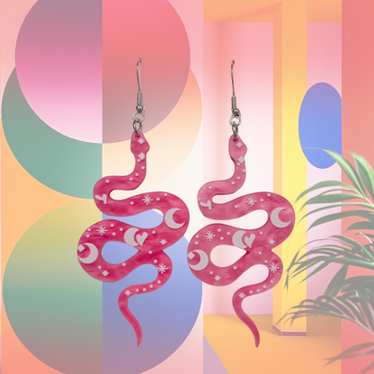 A pair of vibrant pink snake-shaped acrylic earrings featuring whimsical crescent moon and star designs. The lightweight earrings dangle from silver fish hook wires, combining a playful mix of celestial and snake motifs for a bold, statement-making accessory.